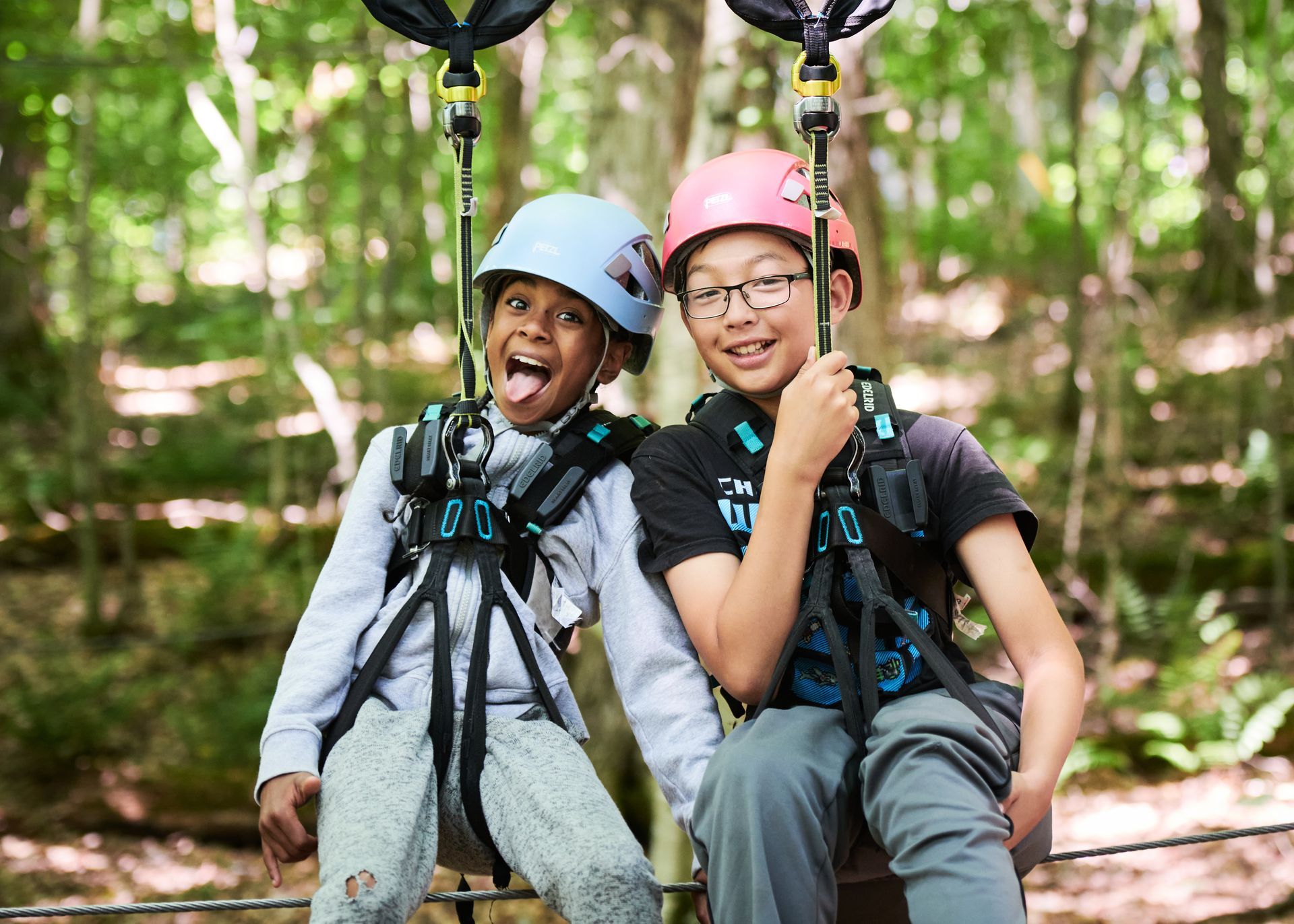 Two children in helmets and harnesses smiling on a zip line course in a forest.