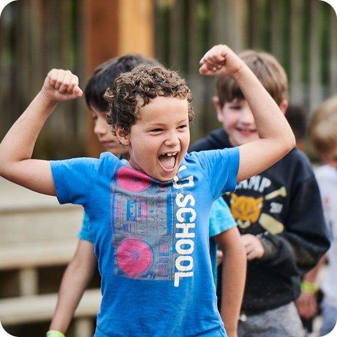 Boy flexing arms, smiling wide at summer camp, others in background.