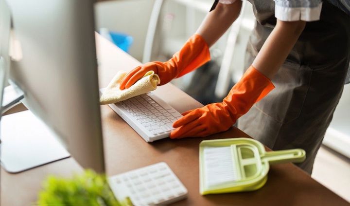 Person wearing orange gloves cleaning a computer keyboard with a yellow cloth on a desk, with a dustpan and brush.