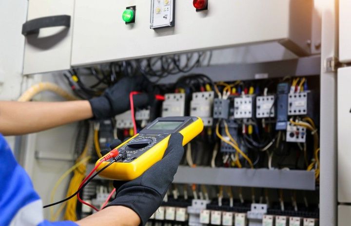 Electrician using a multimeter to inspect wires inside an electrical panel.