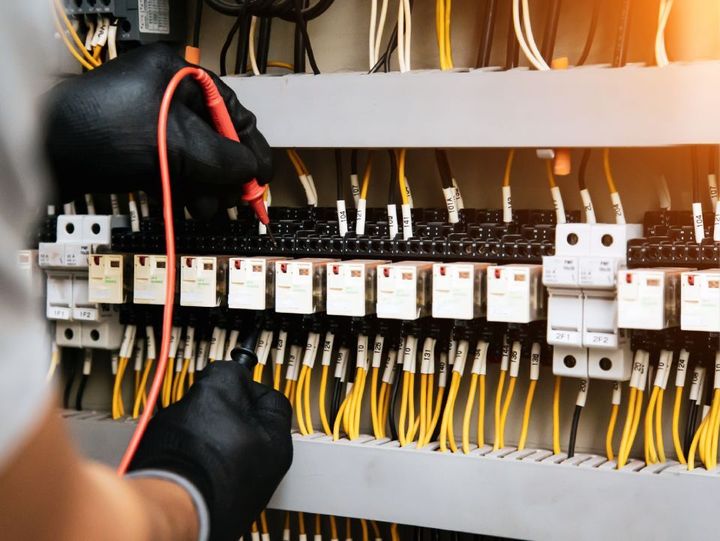 Electrician in black gloves testing wires with a multimeter in a control panel.
