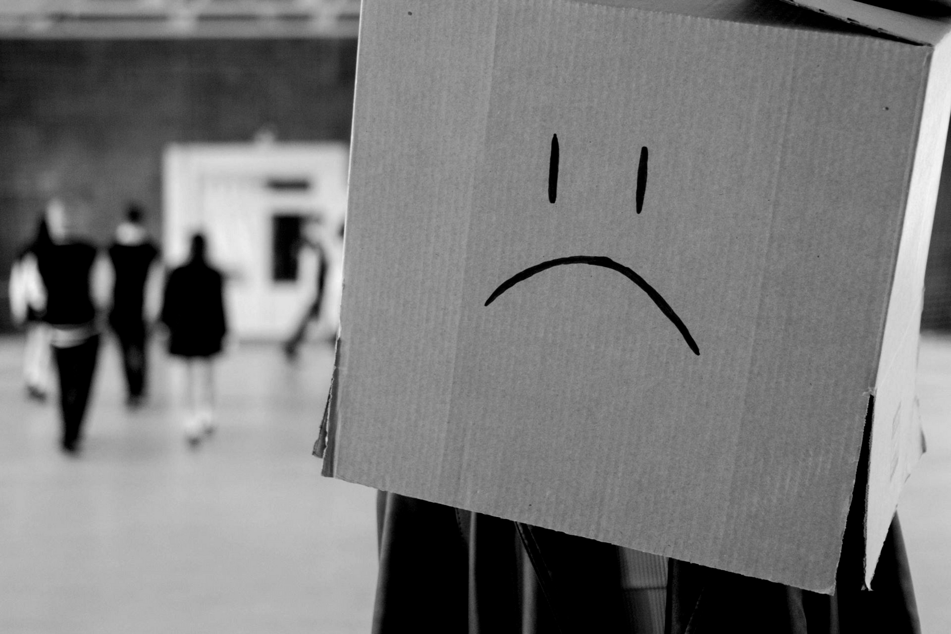 Person wearing a cardboard box with a sad face drawn on it in a hallway