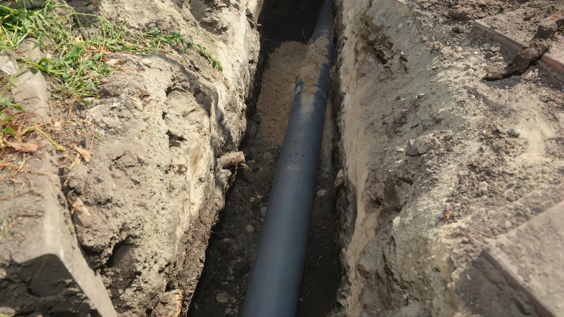 A black pipe lies in a narrow trench filled with loose soil, in an outdoor setting.