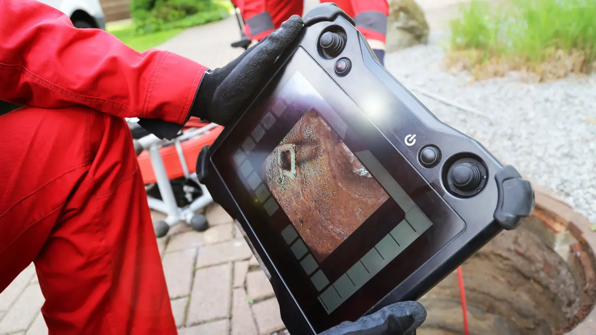 Person in red overalls using a tablet to view the inside of a sewer or manhole, likely for inspection purposes.