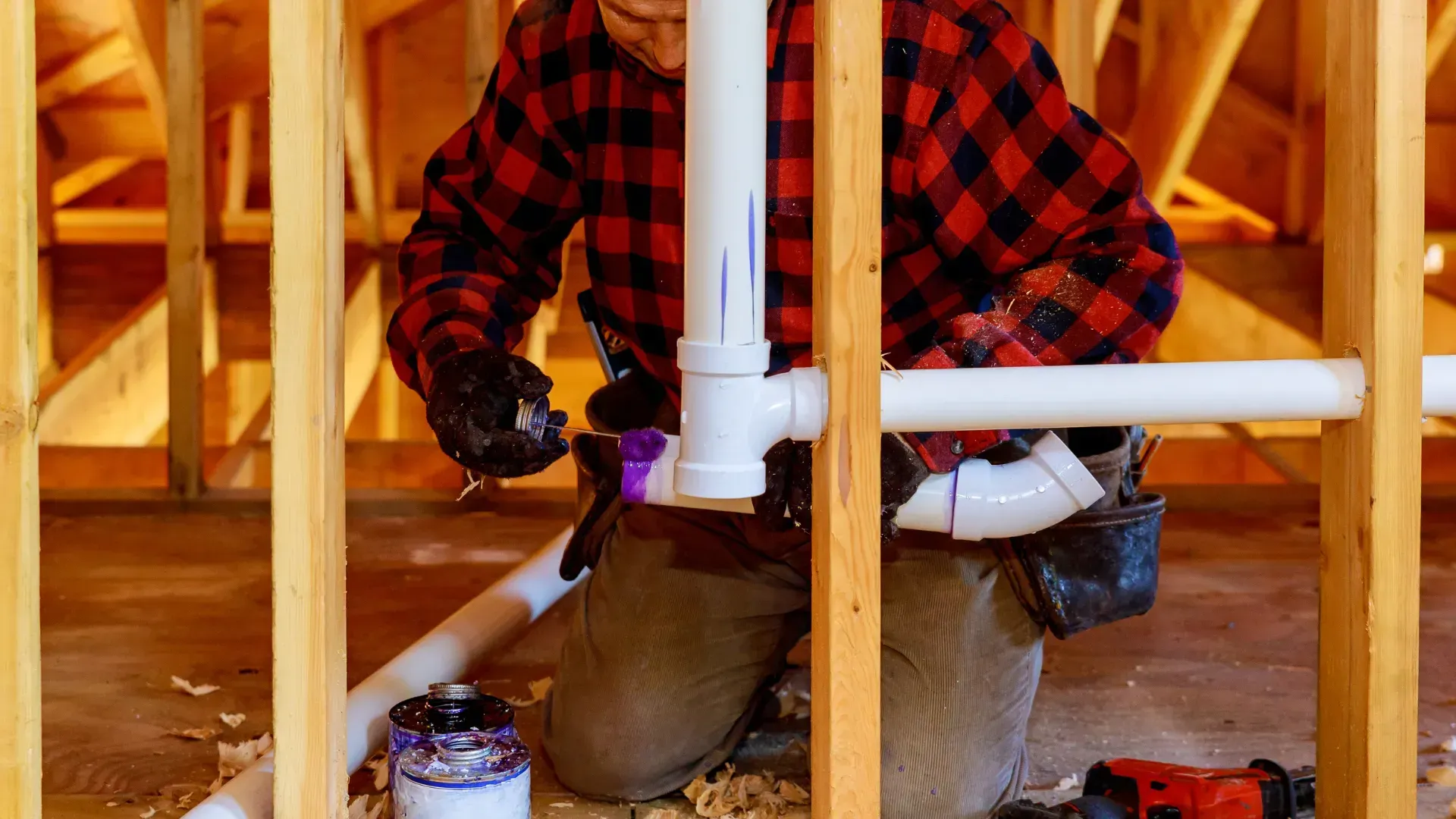 Plumber in a red plaid shirt kneels inside a wooden frame, connecting white PVC pipes.