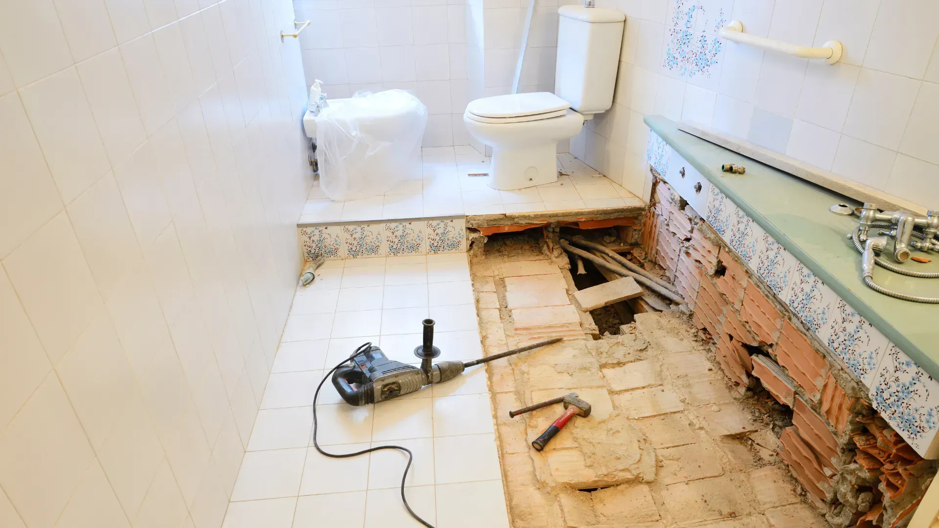 Bathroom under renovation with floor tiles removed, revealing brick and pipes. A drill and tools lie on the floor.