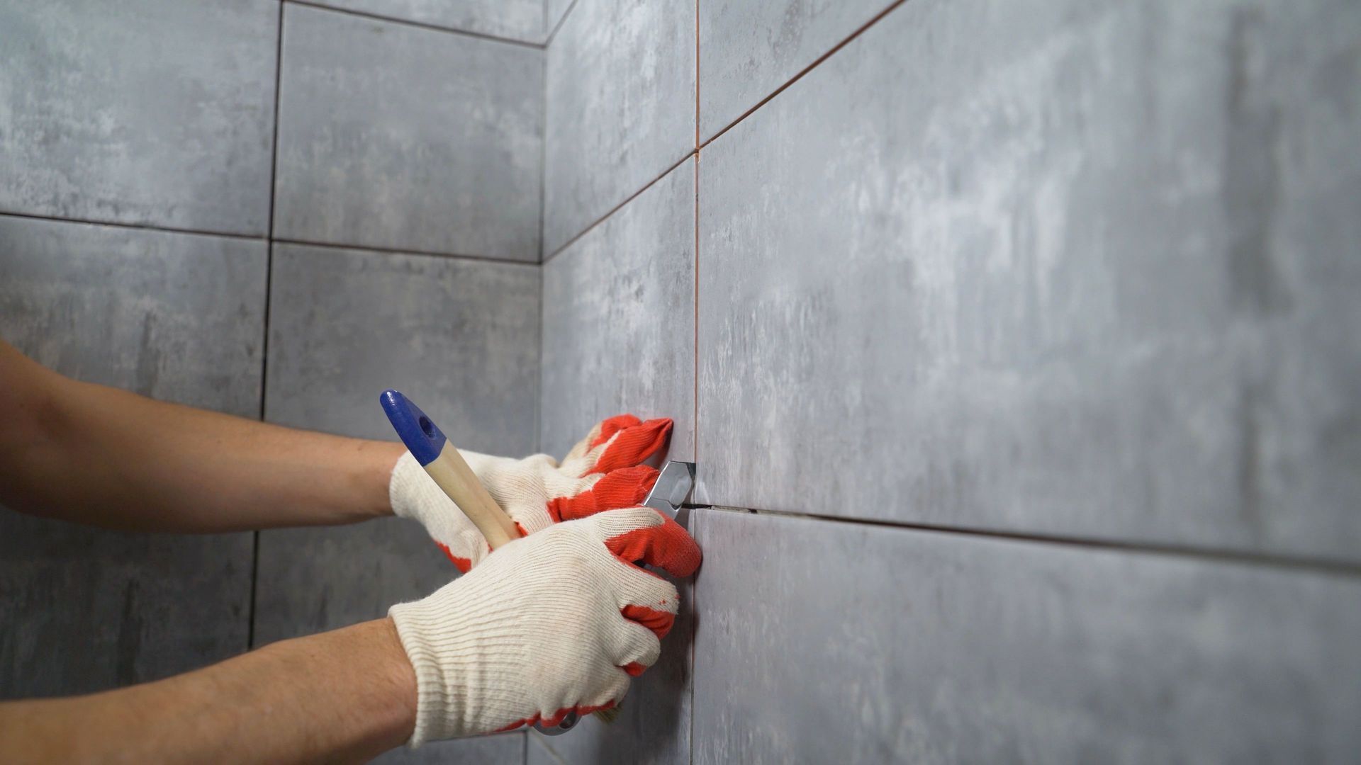 Person wearing gloves uses a tool to work on gray bathroom tile.
