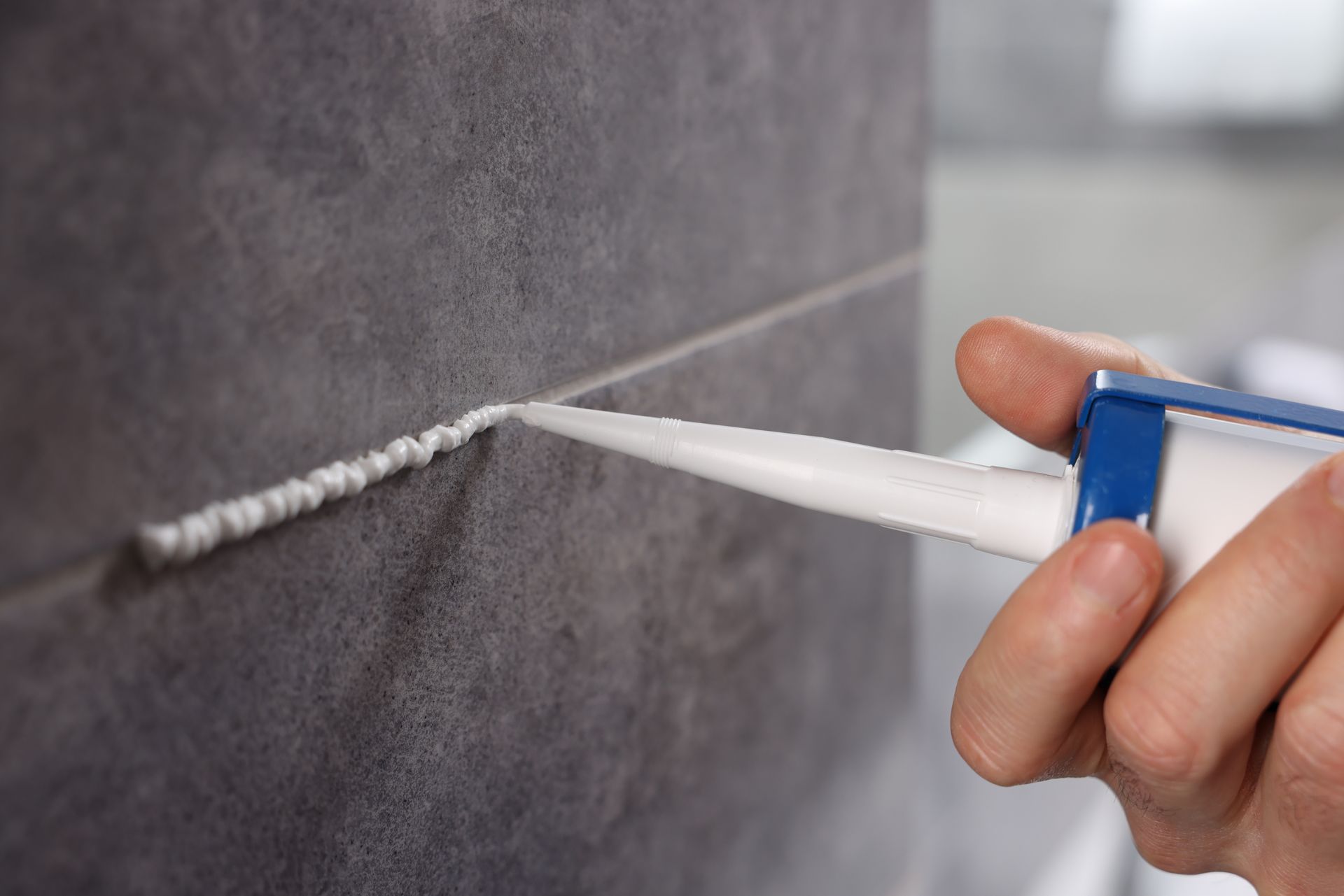 Hand applying white caulk between gray tiles.