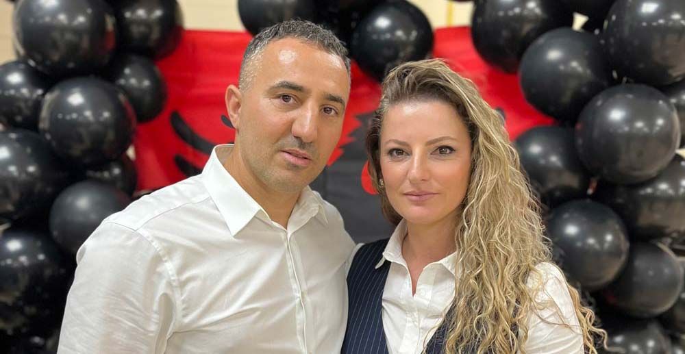 Man and woman posing in front of Albanian flag and black balloons. Both are wearing white shirts.