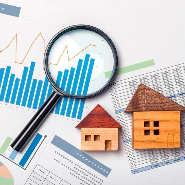 Magnifying glass over graphs and miniature wooden houses; real estate market analysis.