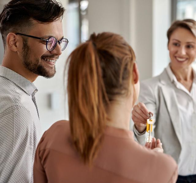 Woman handing keys to a couple; they're smiling. Interior setting.