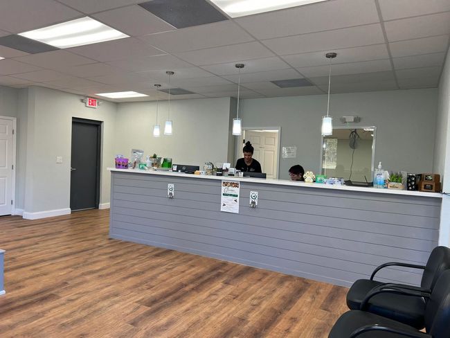 Spacious reception area with long gray counter, wood floor, hanging lights, and black chairs; staff behind desk. Spacious reception area with long gray counter, wood floor, hanging lights, and black chairs; staff behind desk.