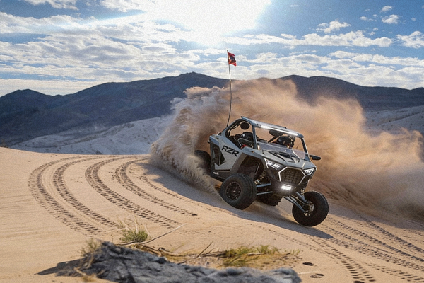 White off-road vehicle kicking up sand, driving fast on a dune. Mountains and sky in the background.