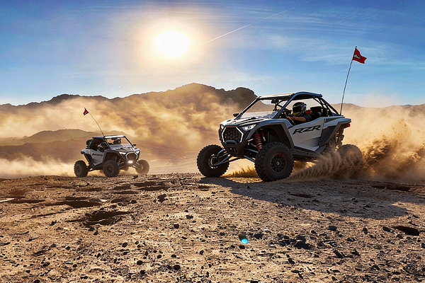 Two off-road vehicles racing across a desert landscape, kicking up dust under a bright sun.
