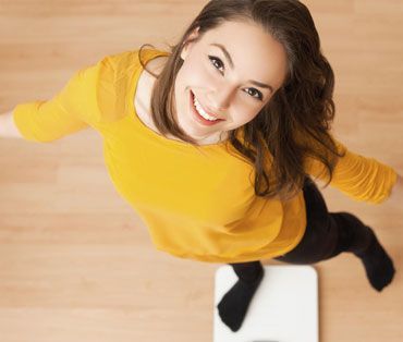 A woman in a yellow shirt is standing on a scale and smiling.