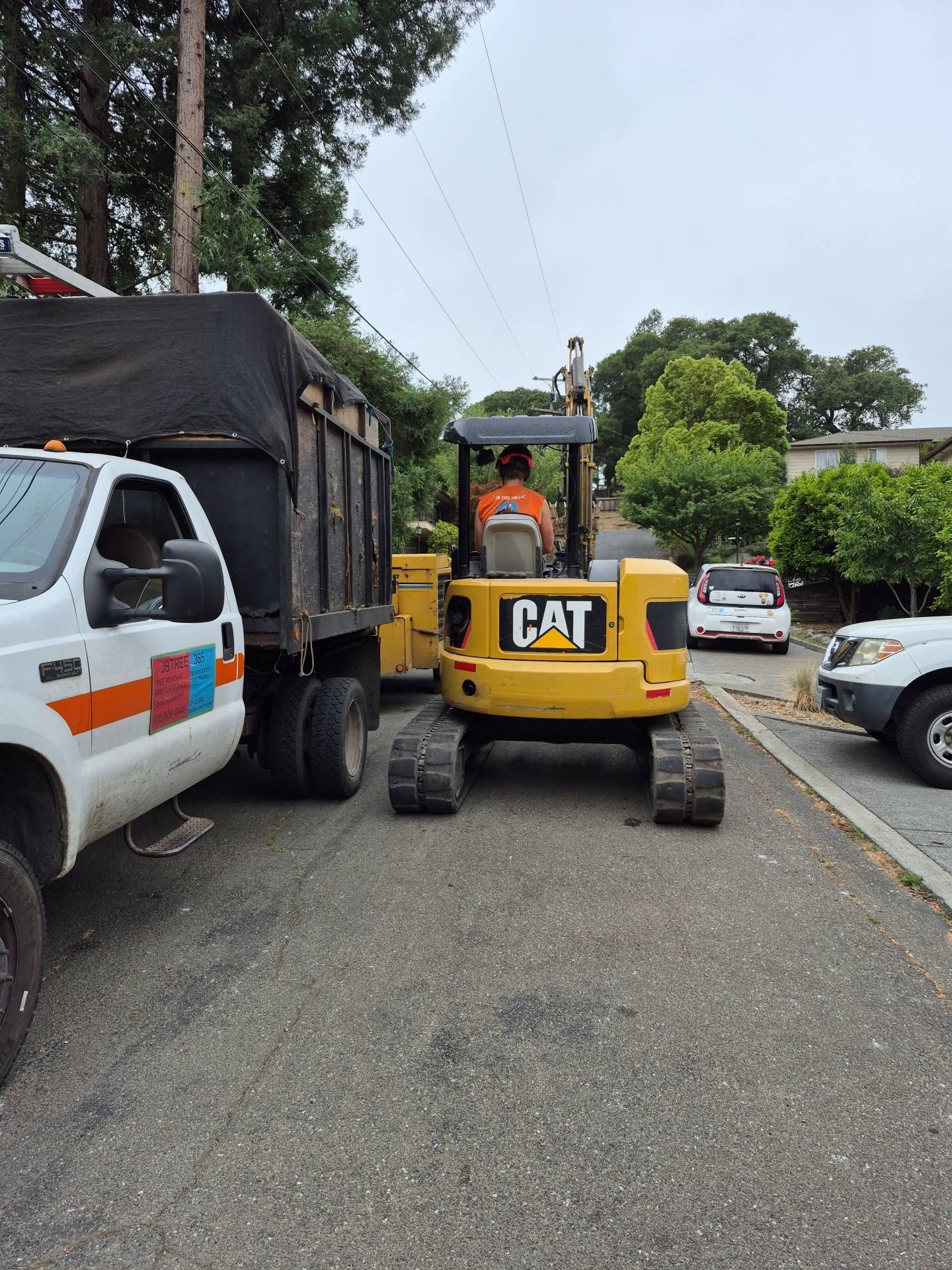 Yellow CAT excavator on a narrow street beside a white truck and parked cars, with trees overhead.