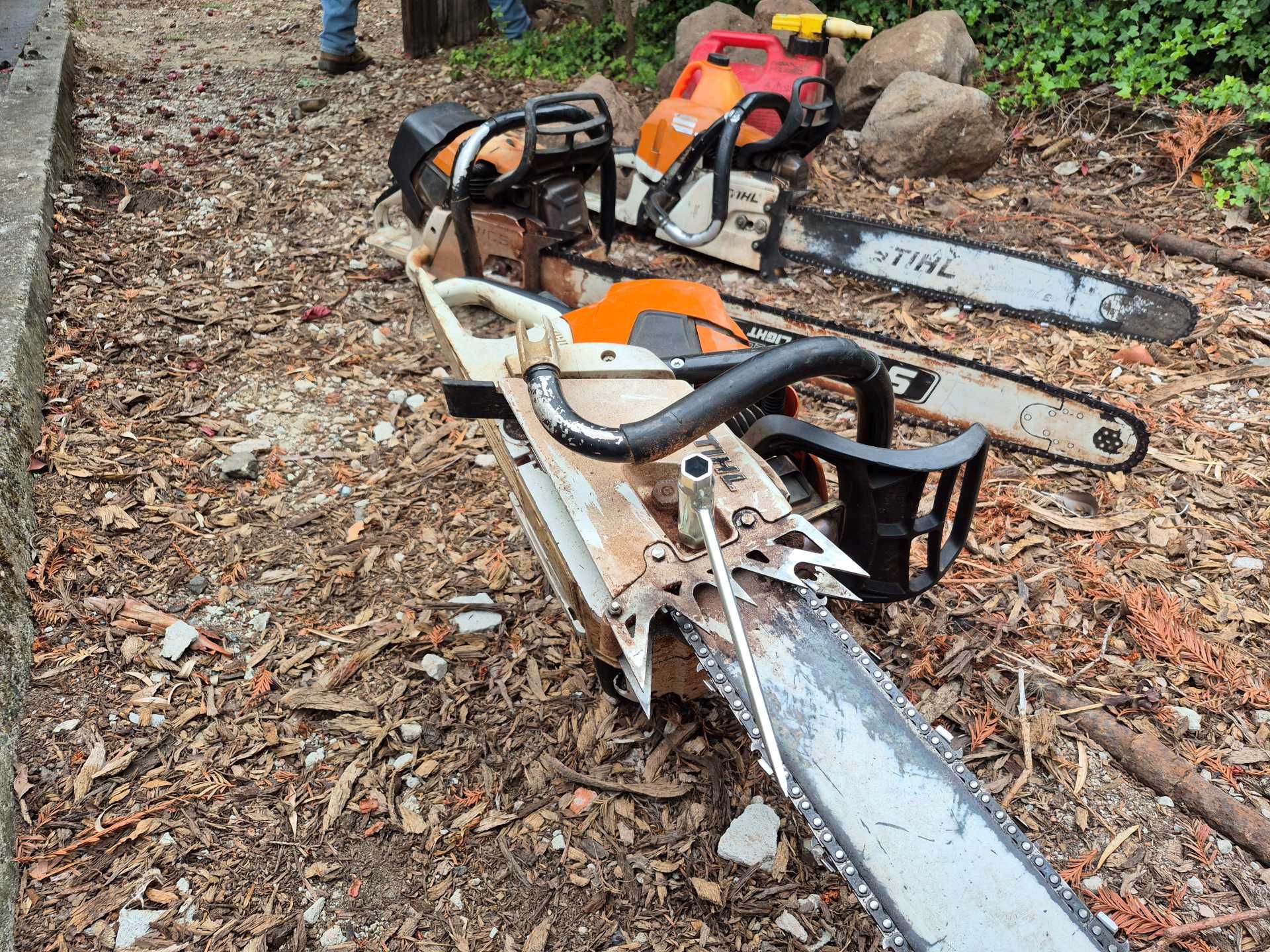 Two chainsaws resting on fallen logs in a wooded area with dry leaves and debris