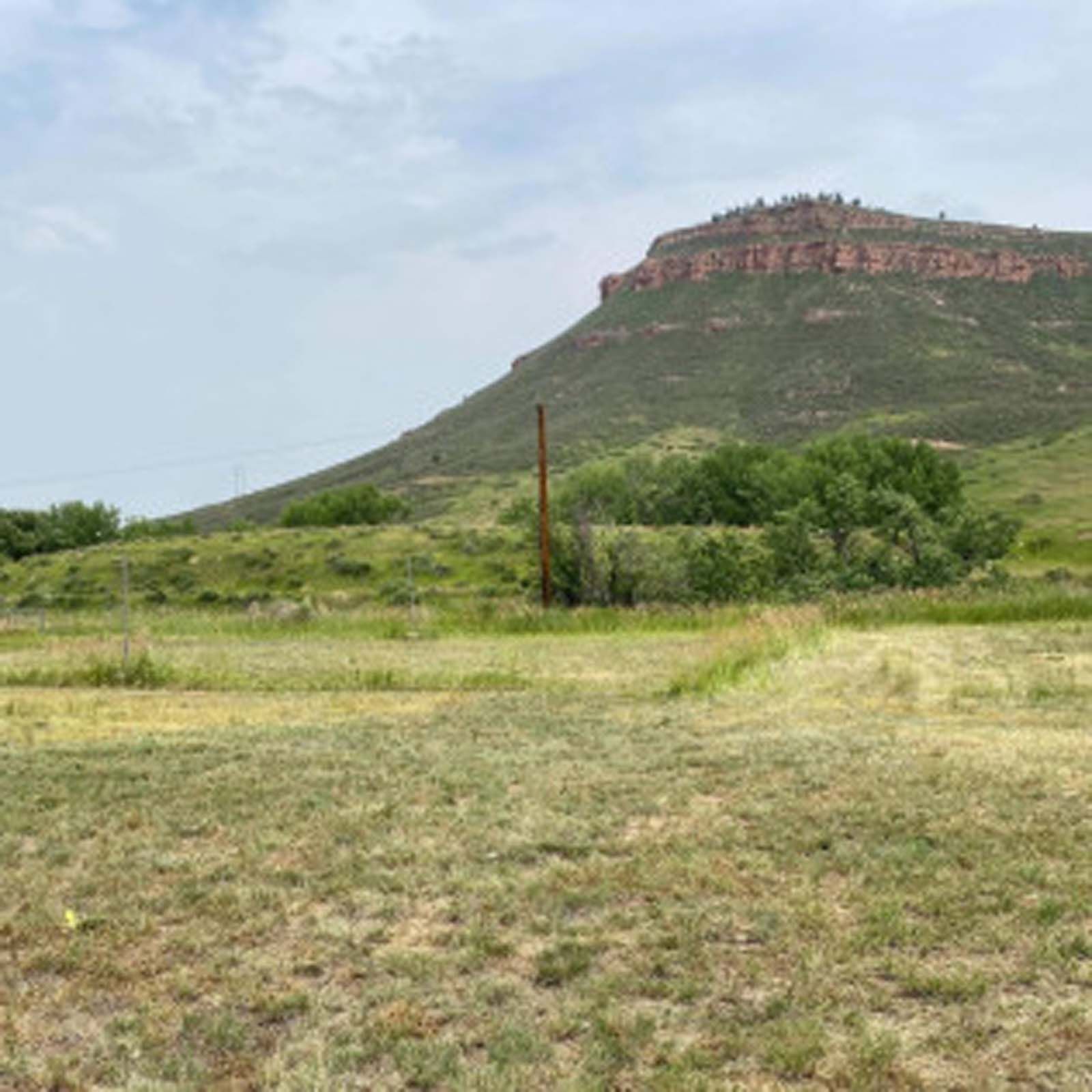 A grassy field leads up to a sloped green hillside topped by a prominent, layered red rock mesa under a cloudy sky.