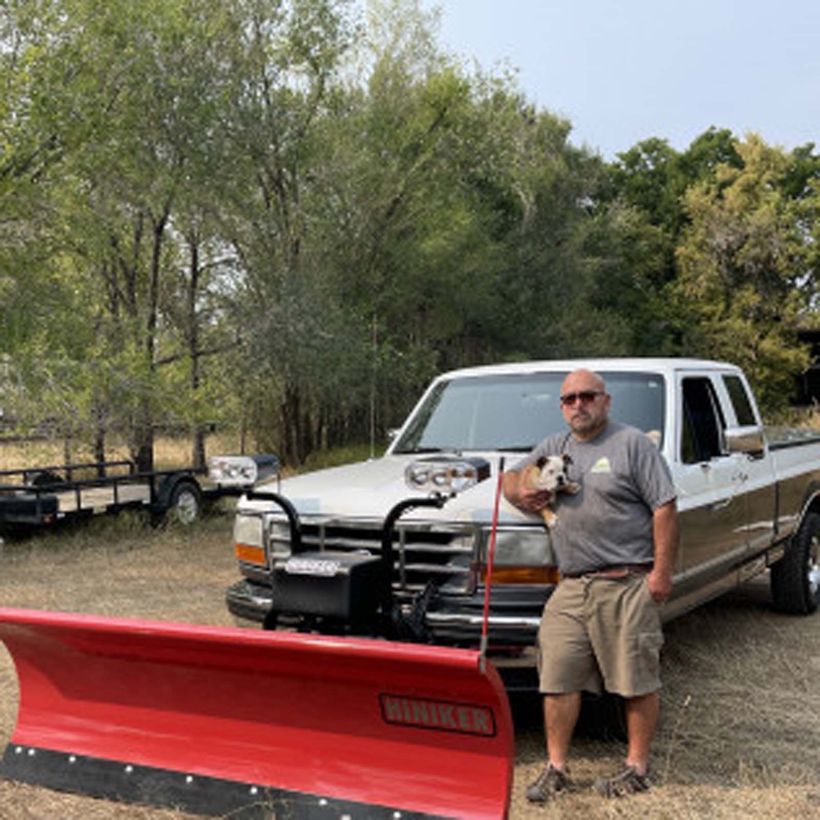 A person standing next to a white pickup truck equipped with a large red Hiniker snow plow, holding a small dog.