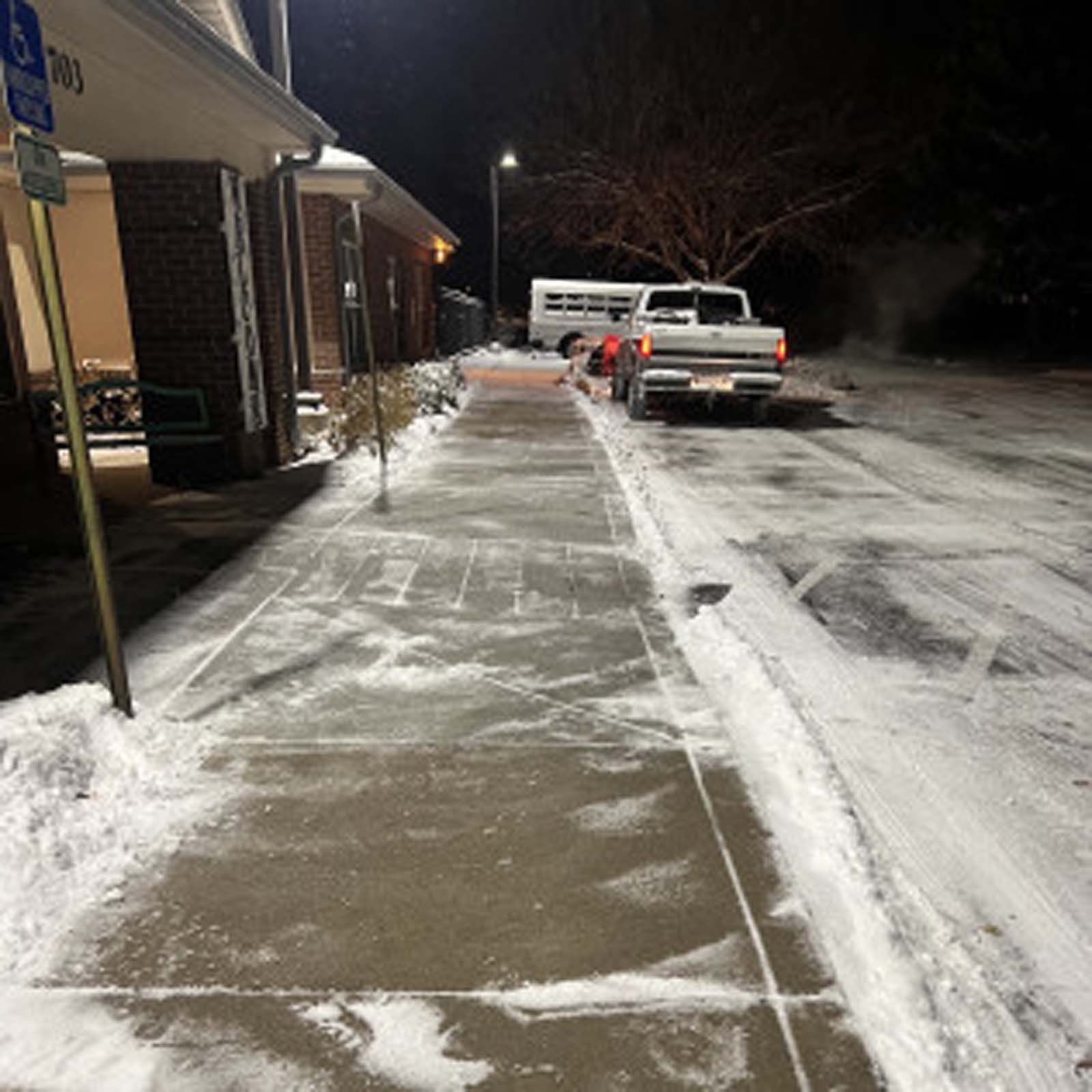 A snowy sidewalk leads toward a building entrance and a parked truck under night lighting.
