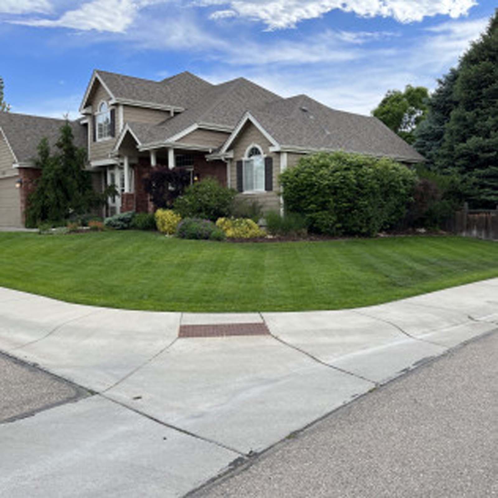 A two-story tan suburban house with a brown roof and a manicured lawn on a corner lot.