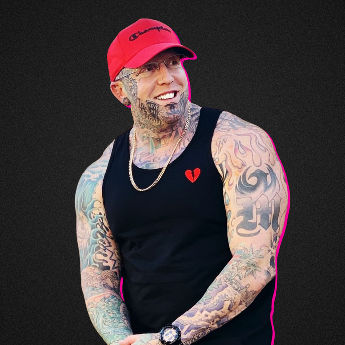 Tattooed person in a red cap and black tank top smiling against a dark background