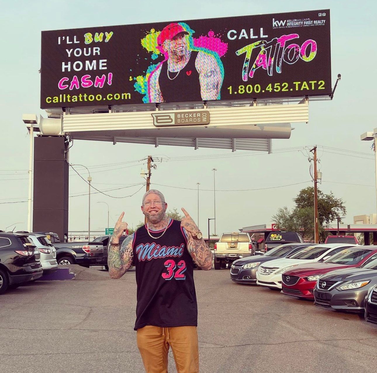Man posing in a car lot beneath a large colorful billboard advertising Call Fatto tattoo shop