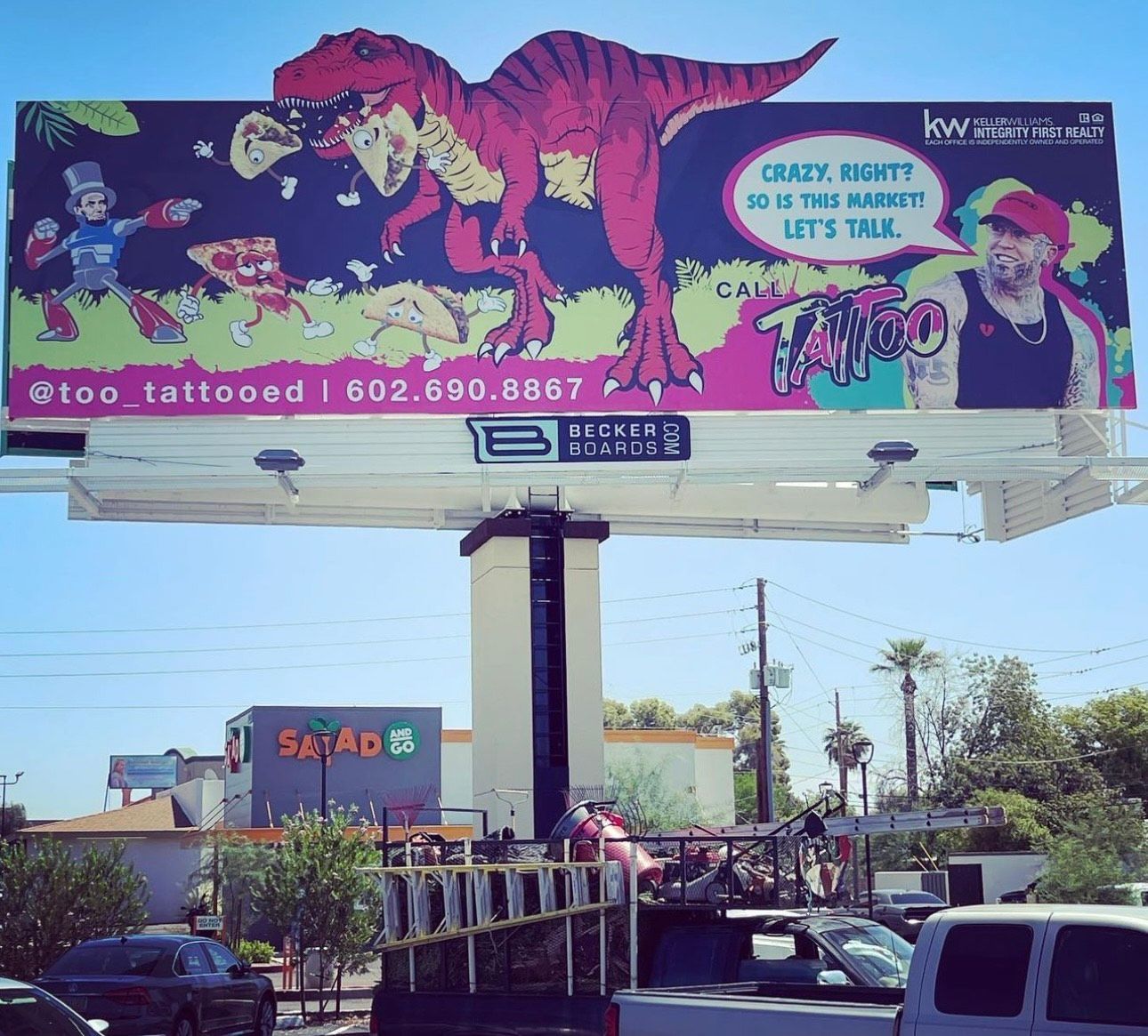 Street billboard with a colorful tattoo ad and dinosaur graphic above a roadside parking lot.