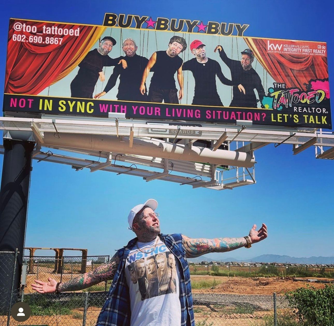 Man standing in a field beneath a colorful roadside billboard, arms outstretched toward the sky.