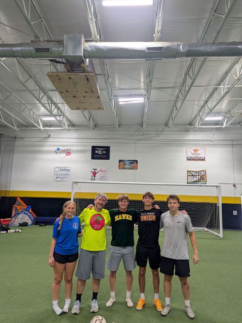 Protect Your Net Trainer/Owner with four players posing in an indoor soccer field in front of a goal, wearing casual athletic clothes.