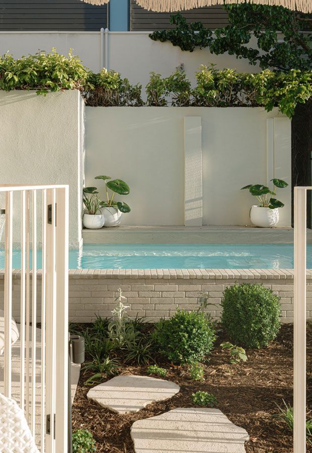 Poolside lounge area with white furniture under a wooden pergola, surrounded by lush greenery.