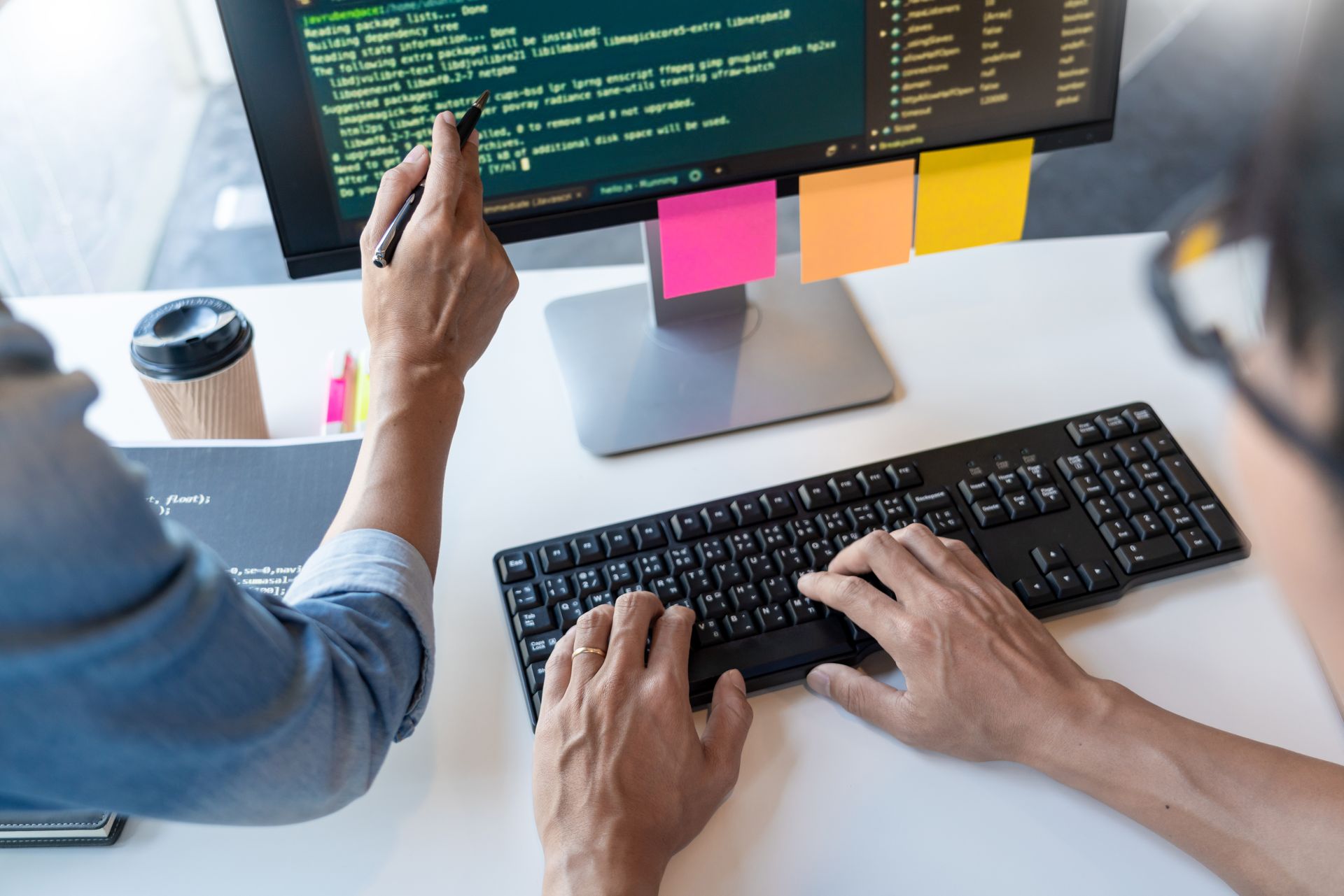 Person typing on a keyboard at a desk with code on a monitor and colorful sticky notes nearby