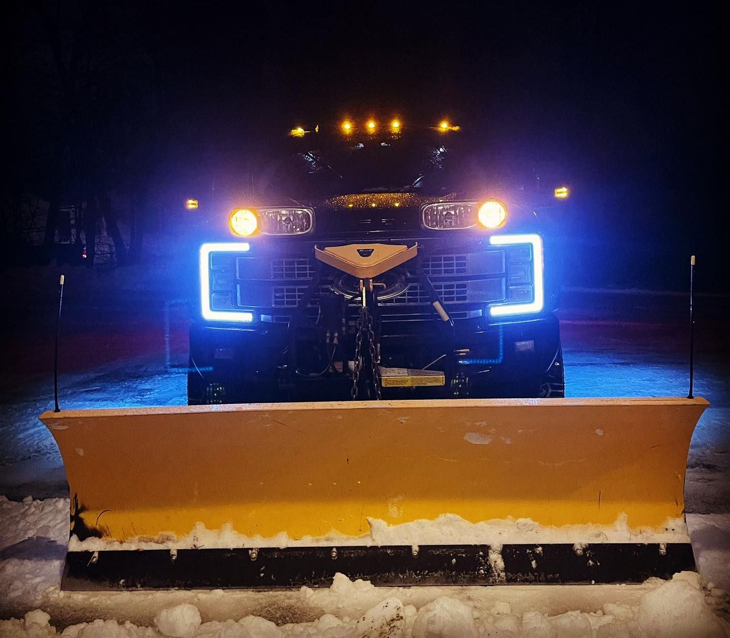 A snow plow is lit up at night in the snow