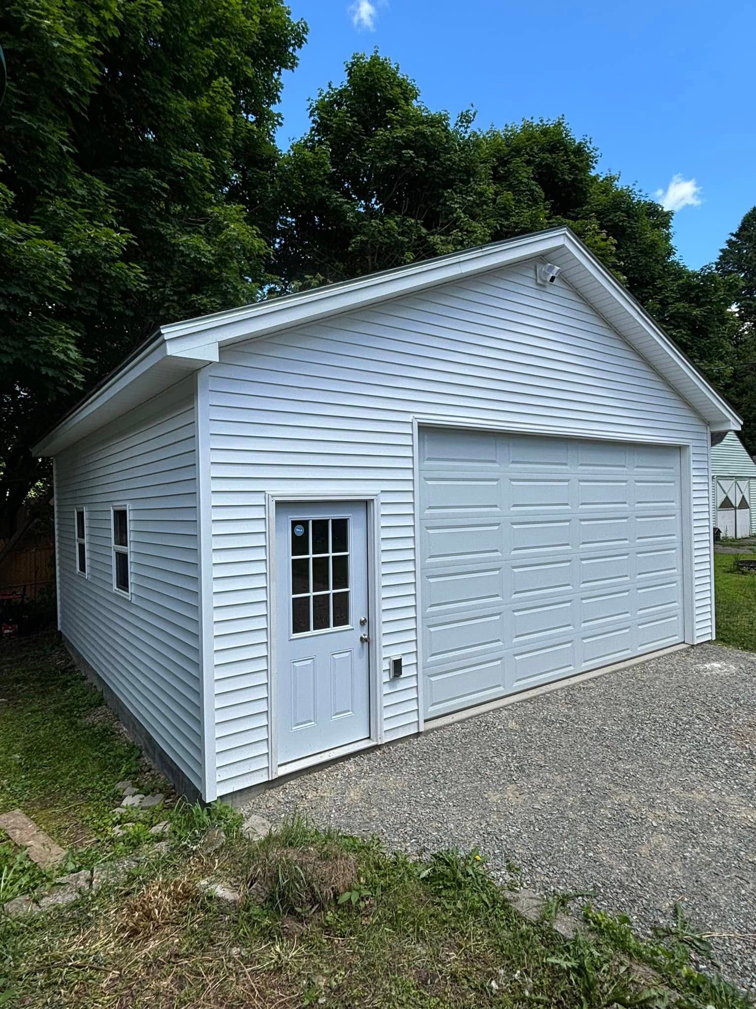 A white garage with a roof and a door is sitting on top of a gravel driveway.