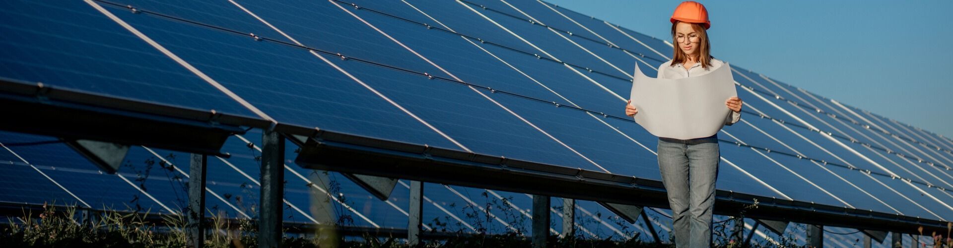 Worker in hard hat holding a laptop in front of solar panels under a blue sky
