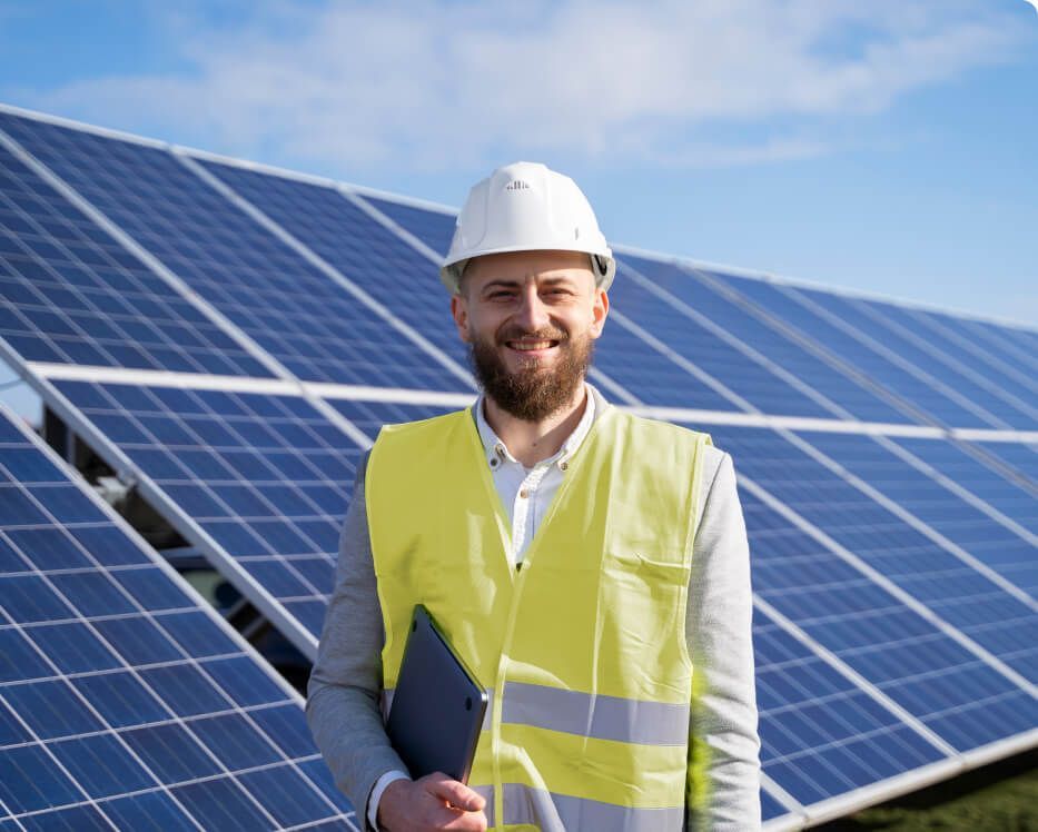 Engineer in white hard hat and yellow vest standing before solar panels, holding a tablet.