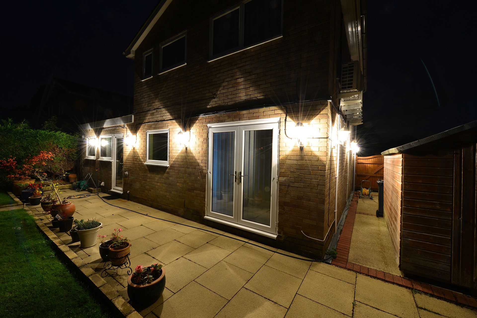 Night-lit brick house patio with glass doors, potted plants, and a side shed along a paved walkway