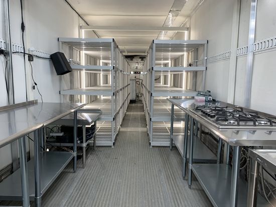 Narrow galley kitchen with stainless steel counters, shelves, and a gas stove in a white trailer-like interior