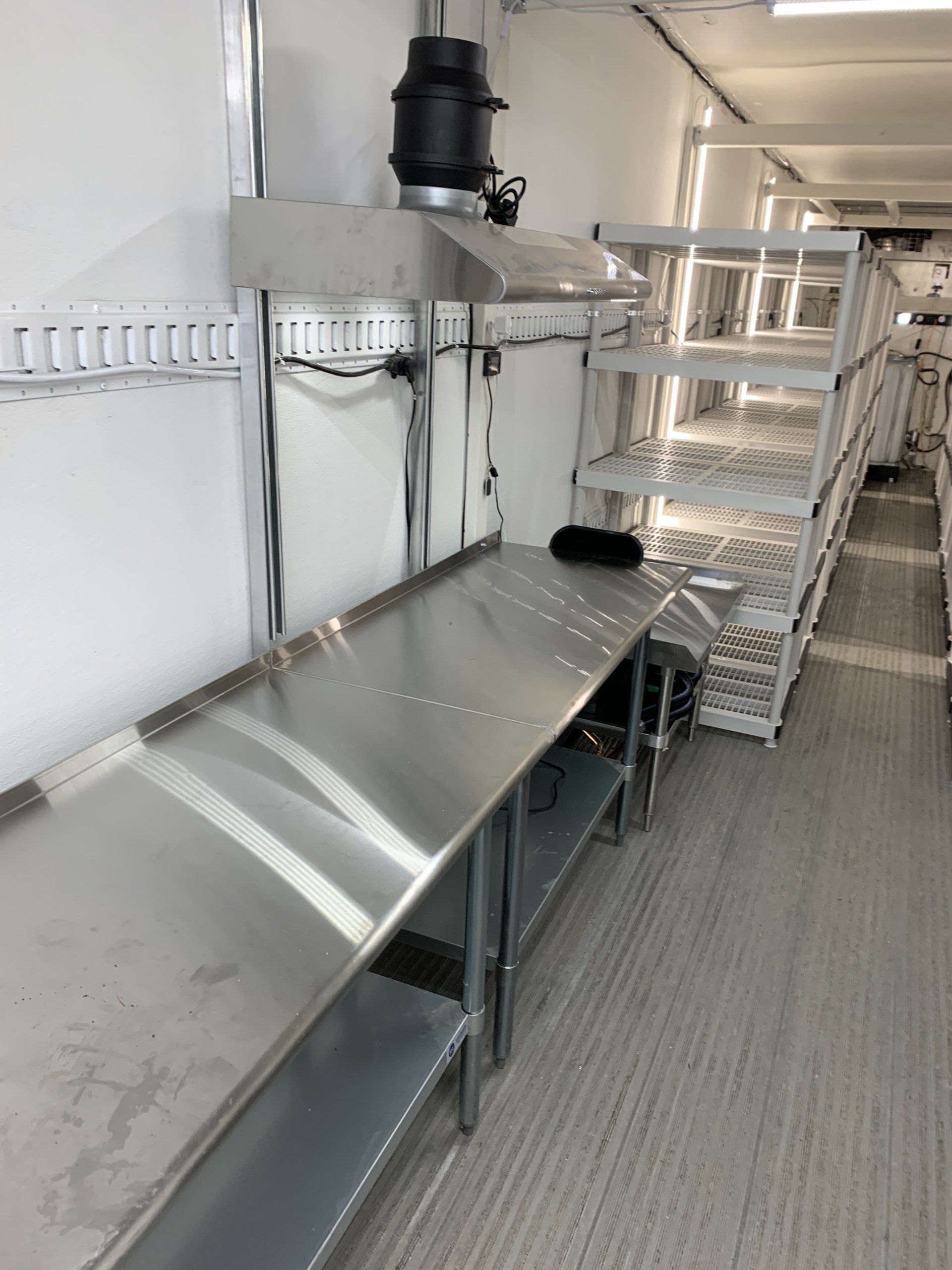 Empty industrial kitchen with stainless steel counters, shelves, and a gray floor ventilation duct.