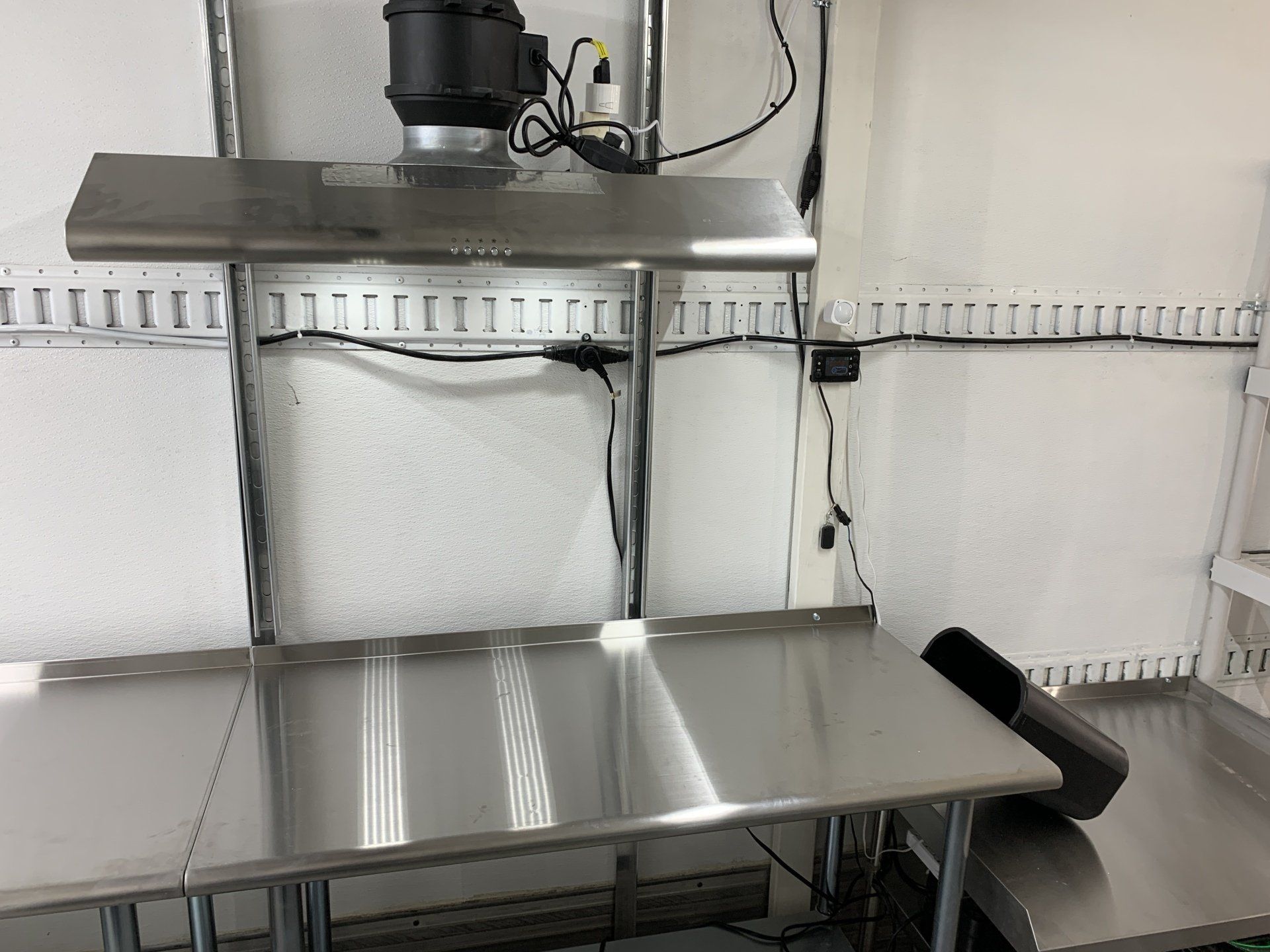 Stainless steel workbench in a lab-like room with shelves, wiring, and a black device mounted above.