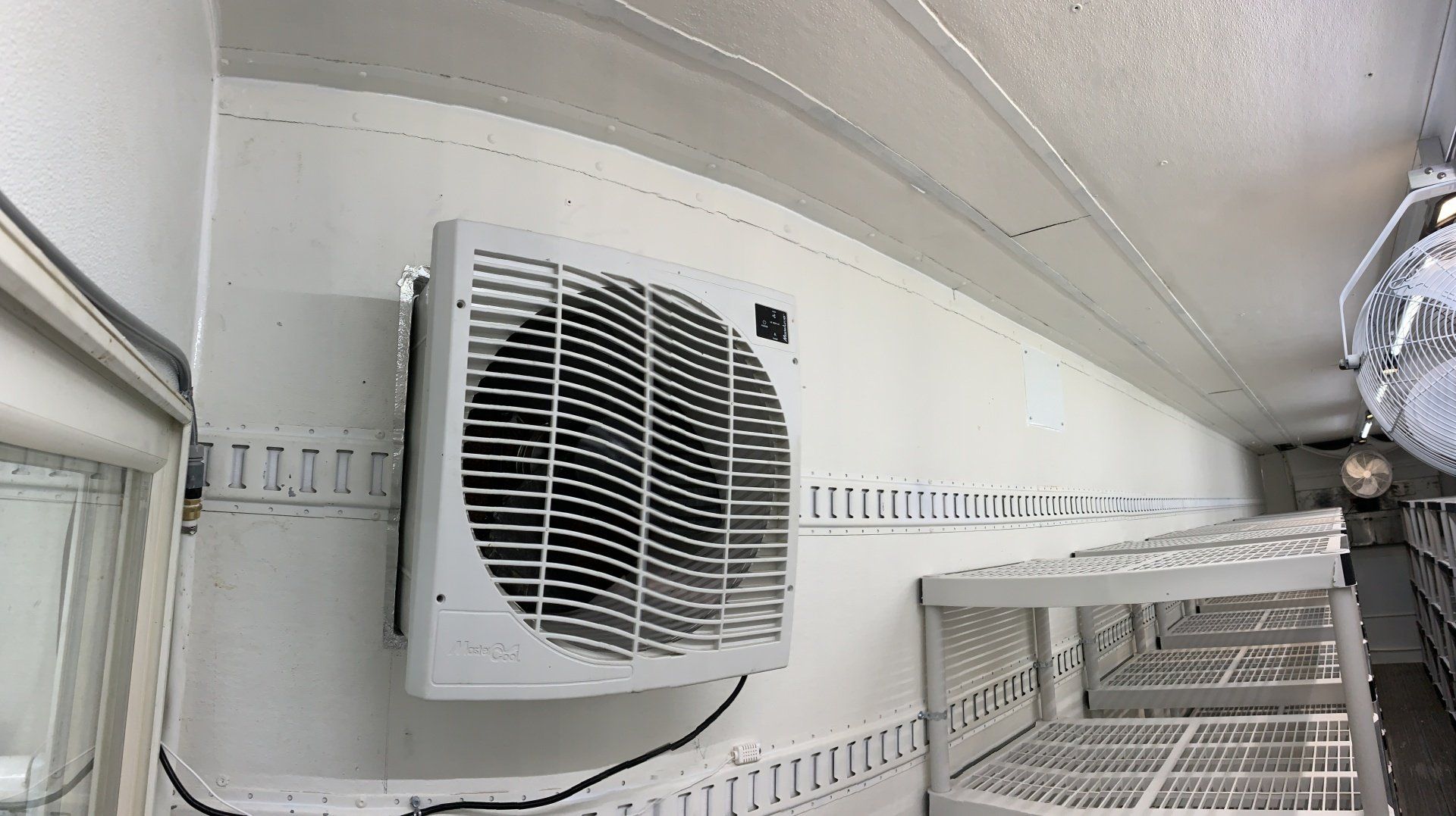 Wall-mounted white ventilation fan in a curved white room, next to metal shelving and ductwork.