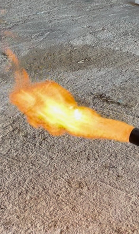 A bright orange flame shooting from a black nozzle against a rough gray ground.