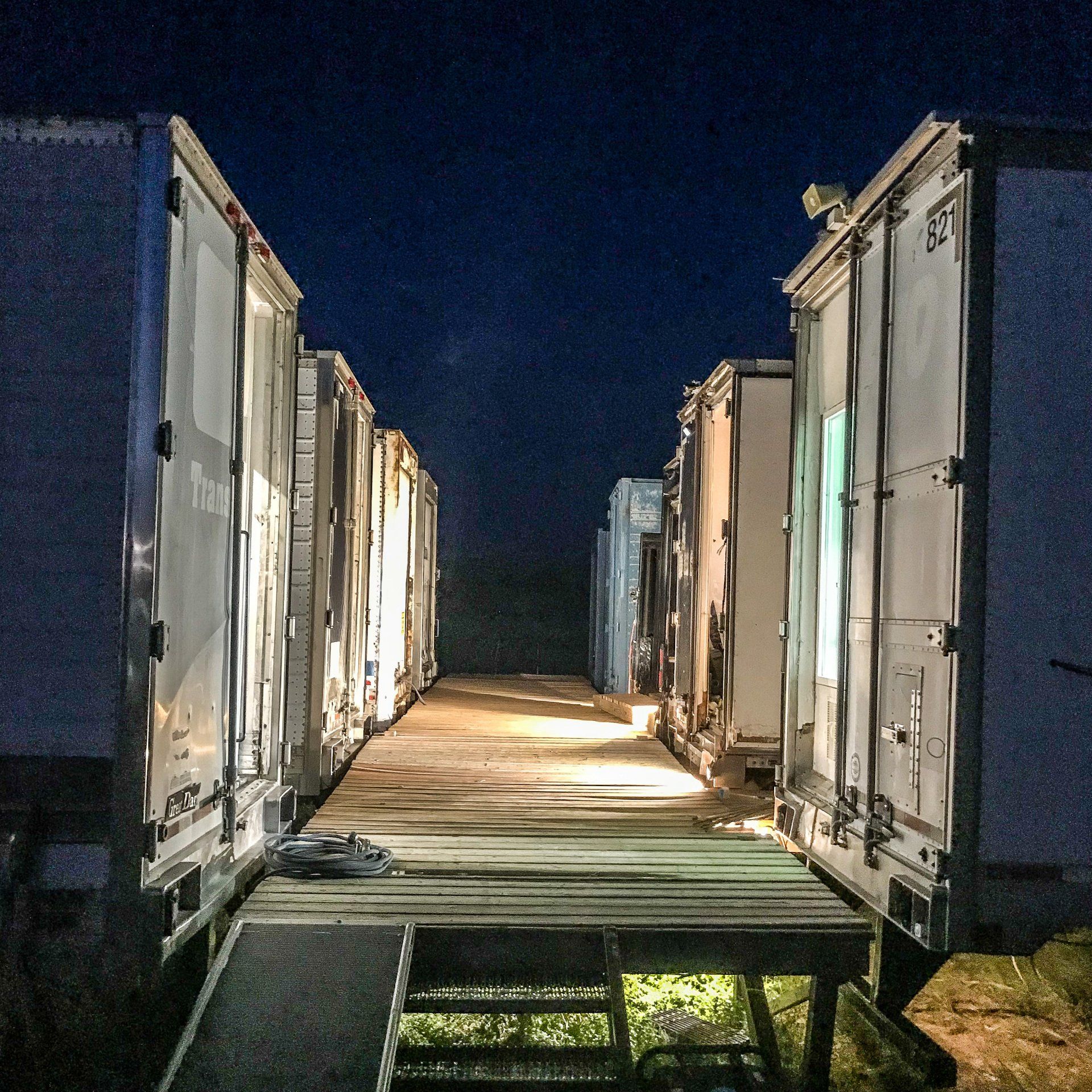 Rows of lit shipping containers at night, forming a narrow walkway between them.