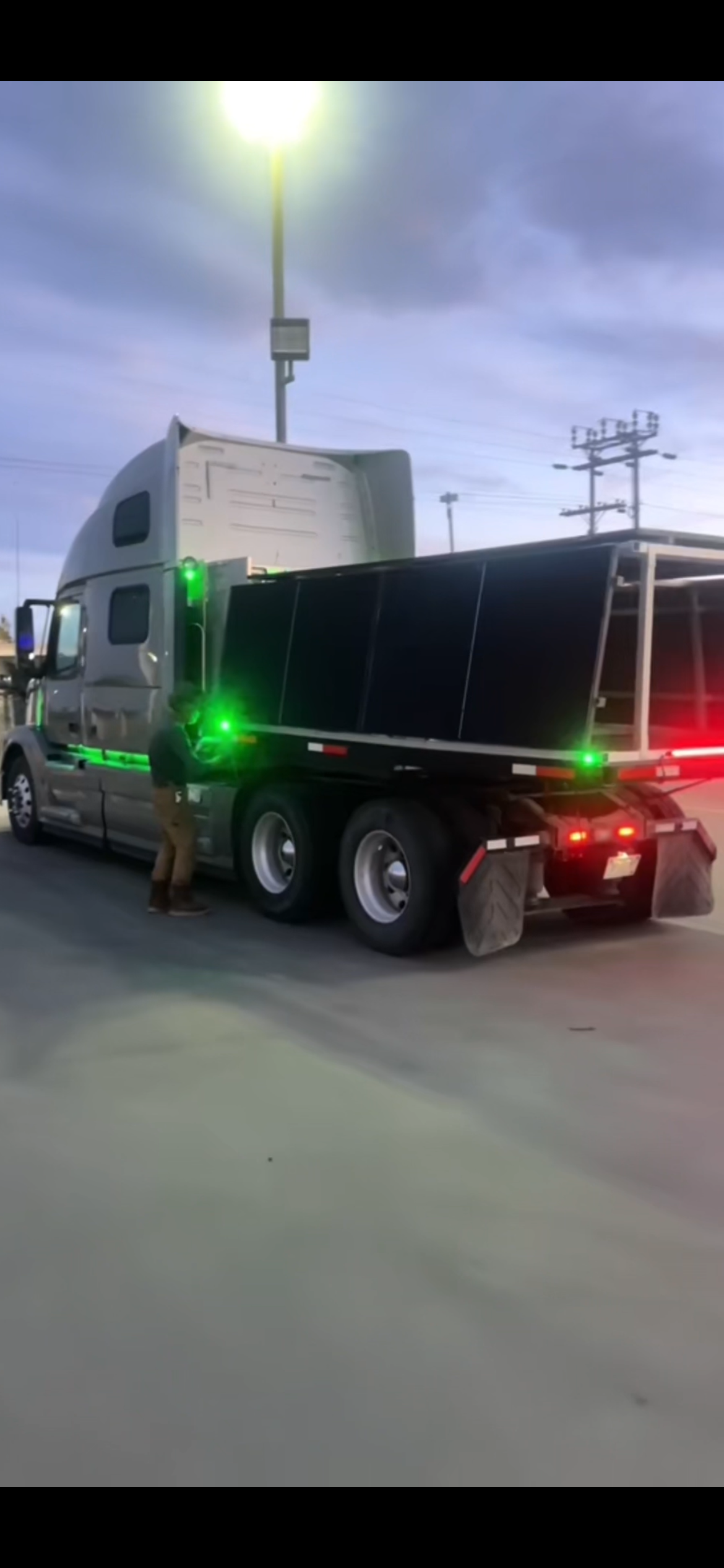 Silver semi-truck hauling a black trailer at dusk in an industrial lot, with red and green lights visible