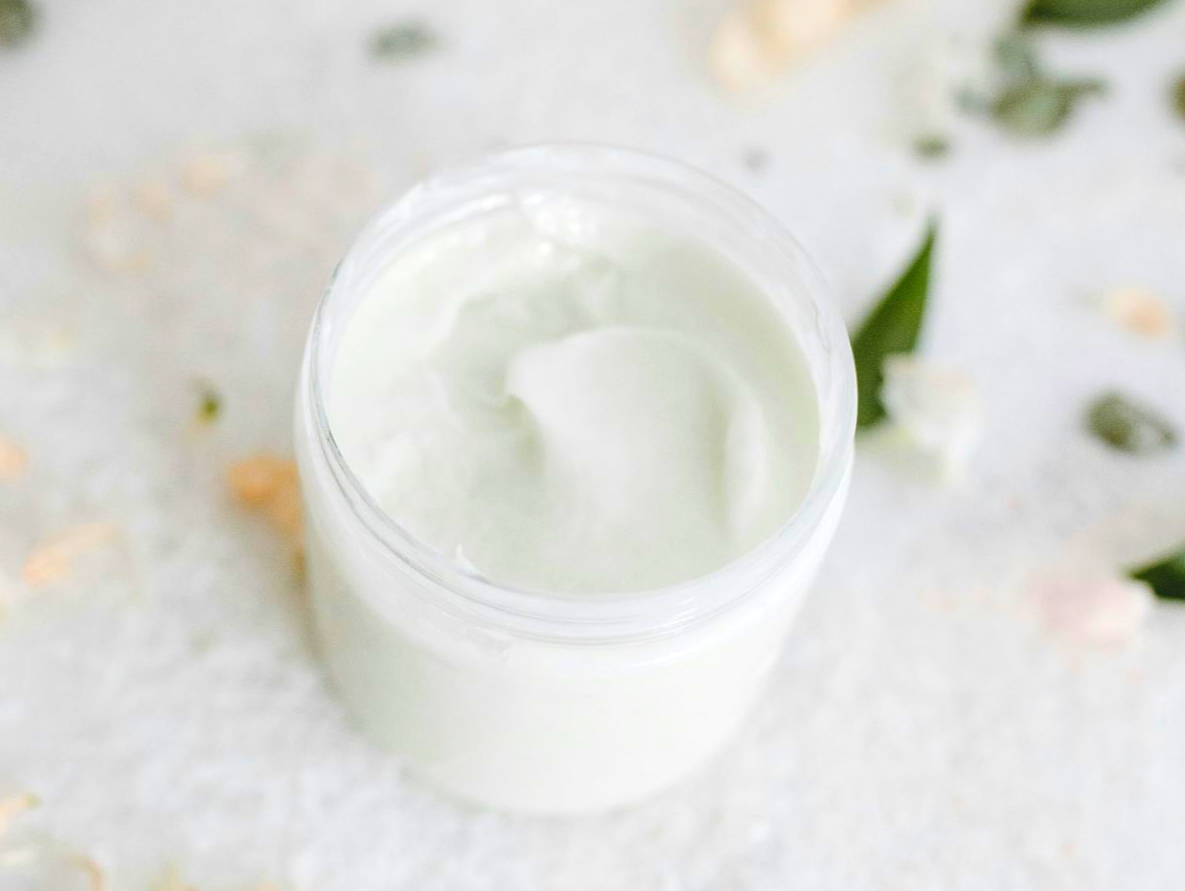 Open jar of white cream on a soft white surface with green leaves nearby