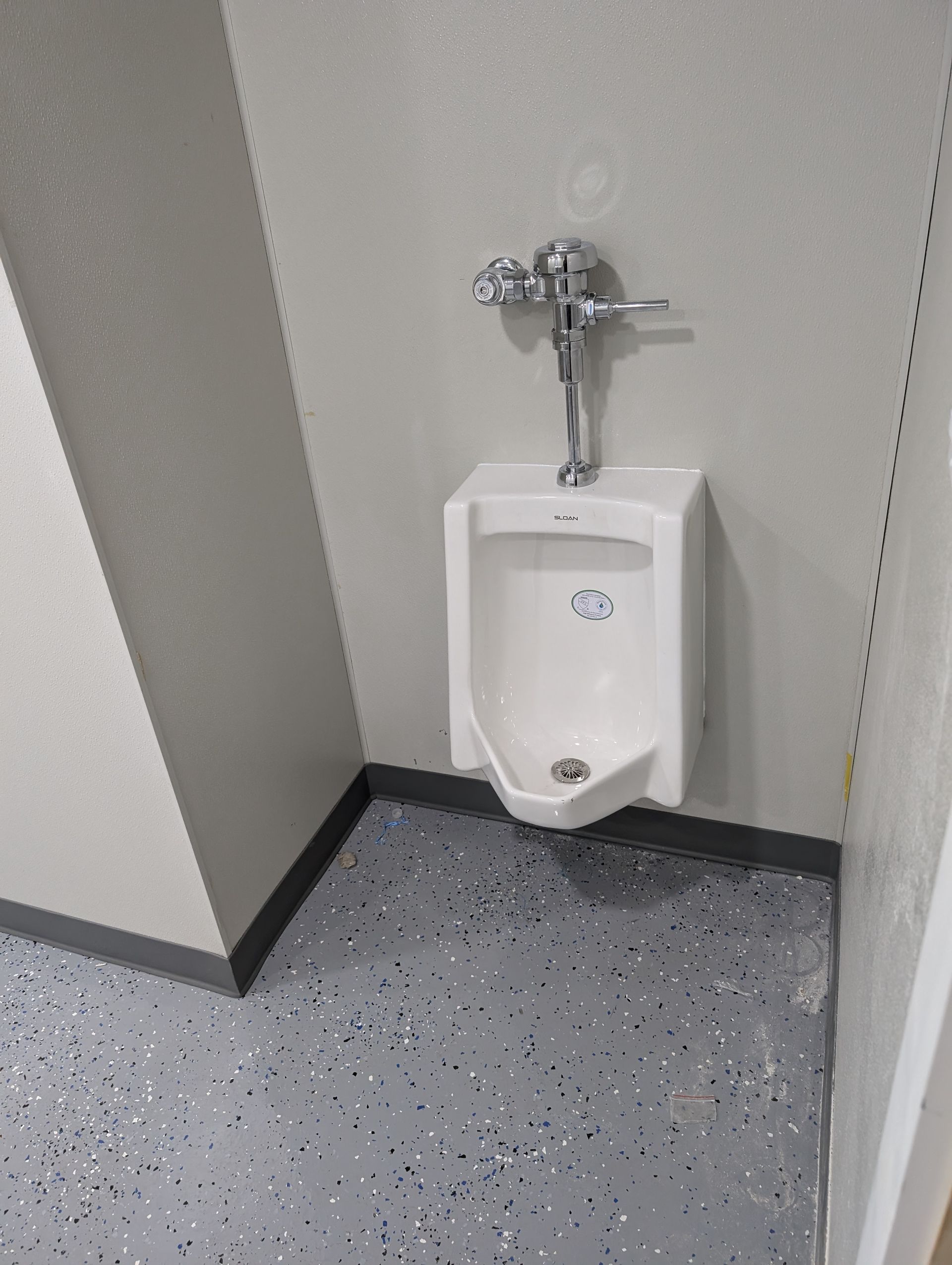 Wall-mounted urinal in a gray restroom corner with speckled floor and black baseboard