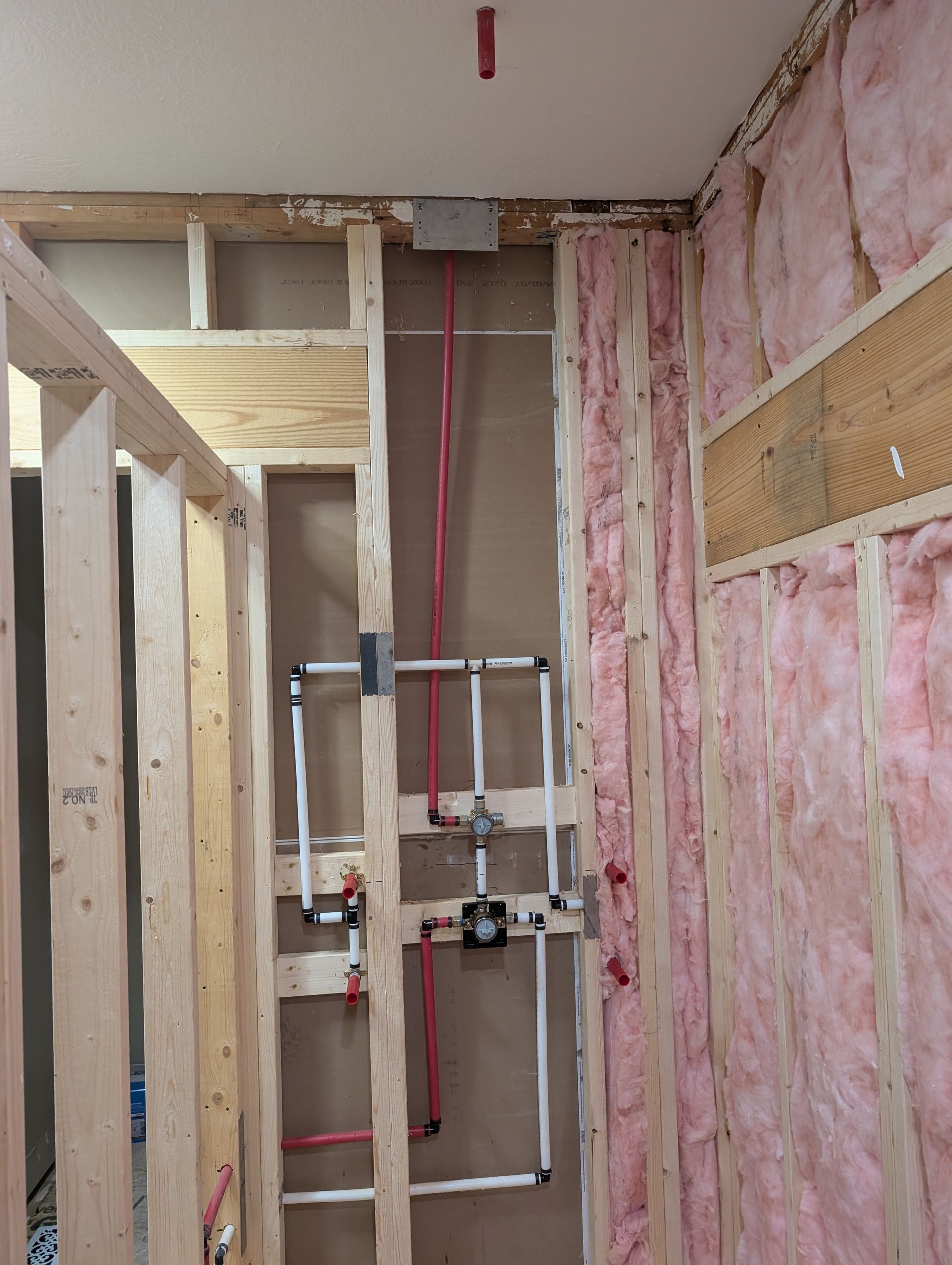 Partially finished bathroom wall with exposed pink insulation and plumbing pipes