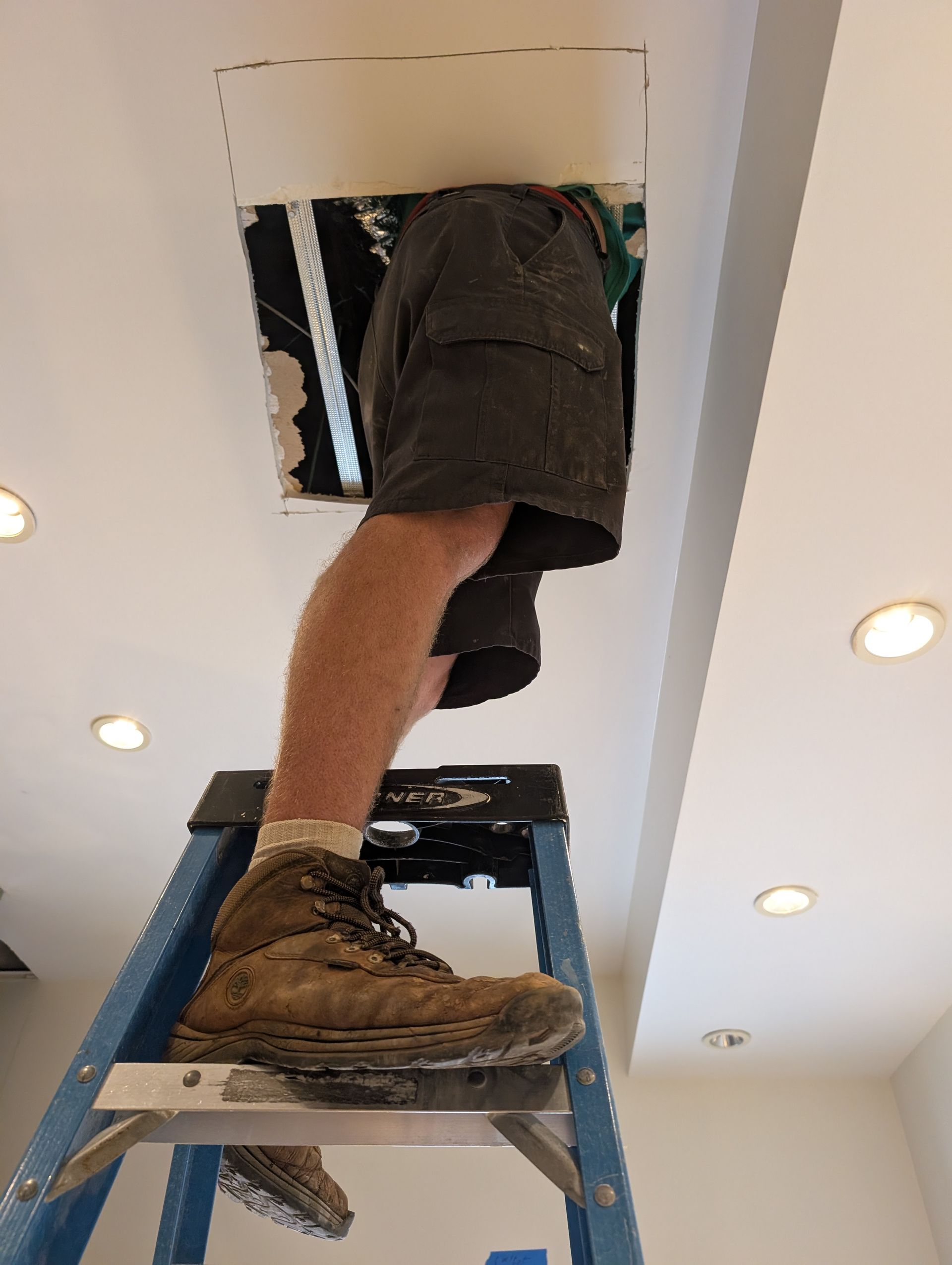 Person on a ladder working in a ceiling attic opening, wearing shorts and work boots.