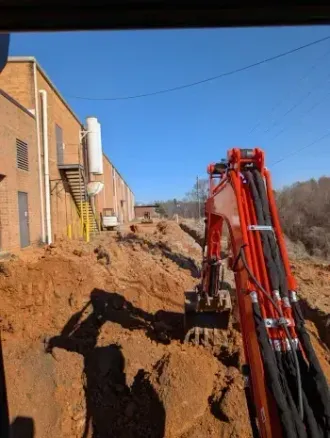 Excavator digging a trench beside a brick building on a dirt construction site under a clear blue sky