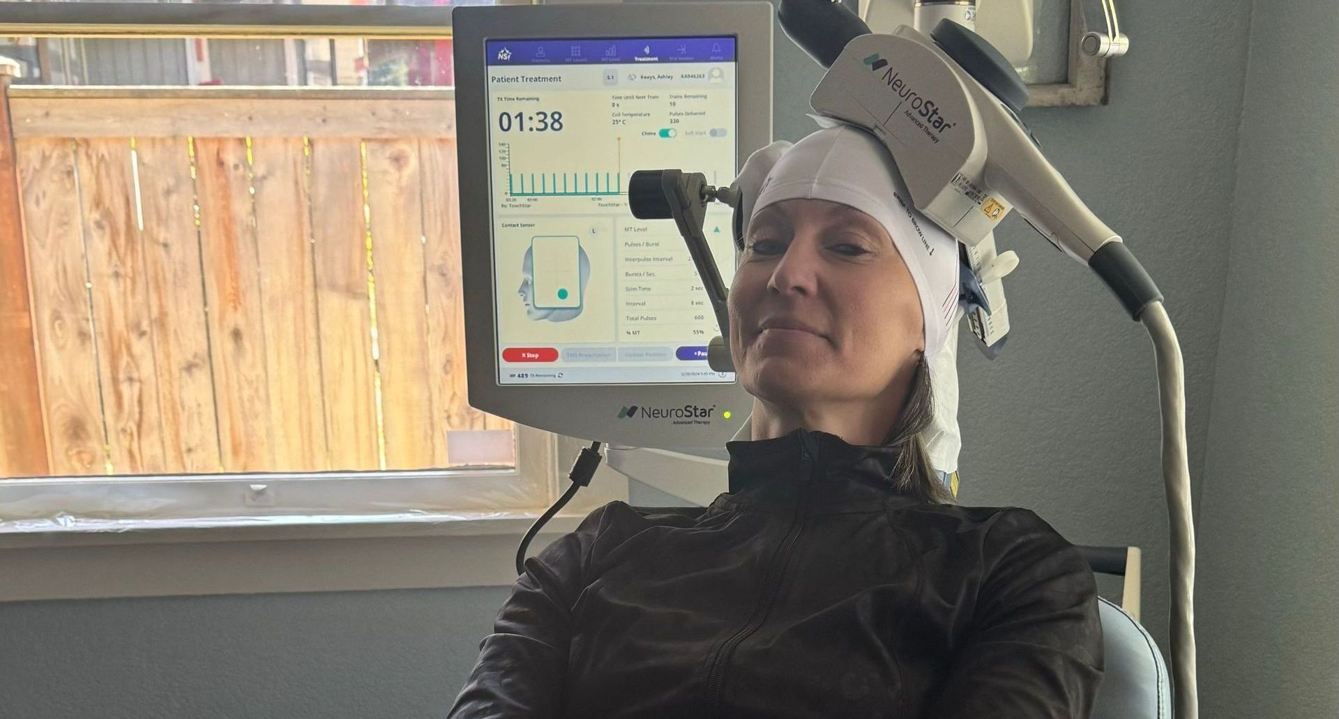 Woman in a gray cap sitting in a clinic chair beside a medical monitor and treatment device.